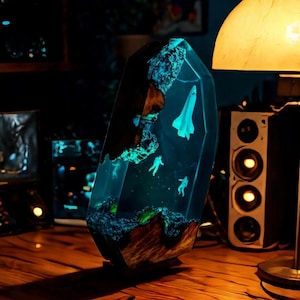 Op de afbeelding: Een decoratieve hars sculptuur met een ruimtethema. De heldere hars bevat een miniatuur spaceshuttle, astronauten en een landschap met gloeiende blauwe en groene accenten. De sculptuur staat op een houten oppervlak.