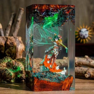 Puede incluir: Escultura de resina con una figura de la serie de anime Attack on Titan. La figura est&aacute; dentro de un bloque de resina transparente con un dise&ntilde;o colorido y en capas. La base es una mezcla de madera y resina, con detalles verdes y naranjas.