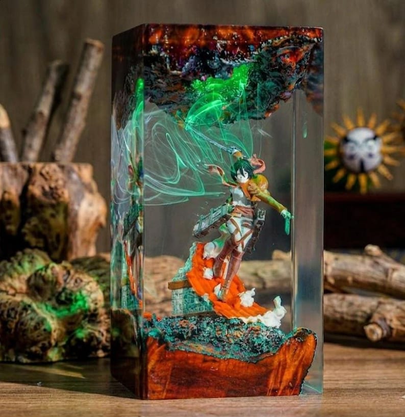 Puede incluir: Escultura de resina con una figura de la serie de anime Attack on Titan. La figura est&aacute; dentro de un bloque de resina transparente con un dise&ntilde;o colorido y en capas. La base es una mezcla de madera y resina, con detalles verdes y naranjas.