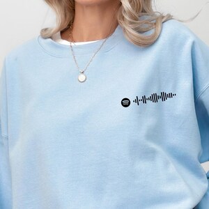 Puede incluir: Sudadera azul claro con un gráfico de código Spotify negro en la parte superior izquierda del pecho. La sudadera es de color liso y tiene cuello redondo. Se ve un collar plateado con un colgante.