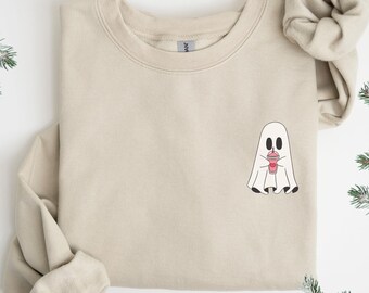 Puede incluir: Sudadera beige con un diseño de fantasma de dibujos animados sosteniendo una bebida rosa. El fantasma tiene ojos y pies negros. La sudadera tiene cuello redondo y mangas largas. El gráfico está en la parte delantera.