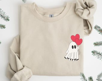 Puede incluir: Sudadera beige con un fantasma de dibujos animados que sostiene dos globos rojos en forma de corazón. El fantasma tiene ojos negros y un cuerpo blanco con dobladillo negro. La sudadera tiene cuello redondo y mangas largas.