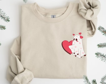 Puede incluir: Una sudadera beige con un fantasma de dibujos animados sosteniendo un corazón rojo. El fantasma es blanco con detalles de corazones rosas. El diseño está en la parte delantera de la sudadera. Las mangas están dobladas. El fondo es blanco con ramitas verdes.