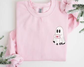 Puede incluir: Sudadera rosa claro con un fantasma de dibujos animados sosteniendo una carta con un corazón. El fantasma tiene ojos y pies negros. La sudadera es de color liso con cuello redondo y mangas largas. El diseño está en la parte inferior derecha.