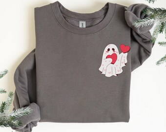 Puede incluir: Una sudadera gris oscuro con un gráfico de fantasma sosteniendo un corazón rojo. El fantasma es blanco con detalles de corazones rosas. La sudadera está doblada, con las mangas bien metidas.