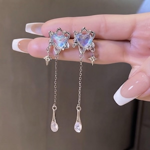 May include: A pair of silver-toned drop earrings featuring a central iridescent heart-shaped stone encased in a star-shaped frame. Each earring includes a dangling silver chain ending in a teardrop-shaped crystal charm and a small star accent.