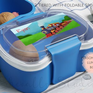 May include: Blue two-tiered lunch box with a clear lid and a foldable spoon. The lid features a personalized label with a cartoon train and the name "Ryan". The lunch box is labeled "Personalized Lunch Box".