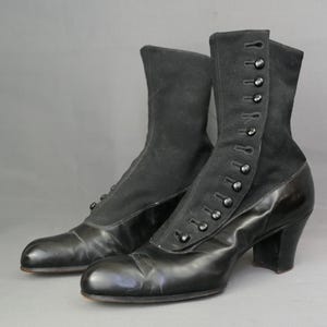 May include: A pair of black vintage ankle boots. The boots have a combination of smooth leather and suede-like fabric. They feature button closures and a small heel. The boots are likely from the Victorian or Edwardian era.