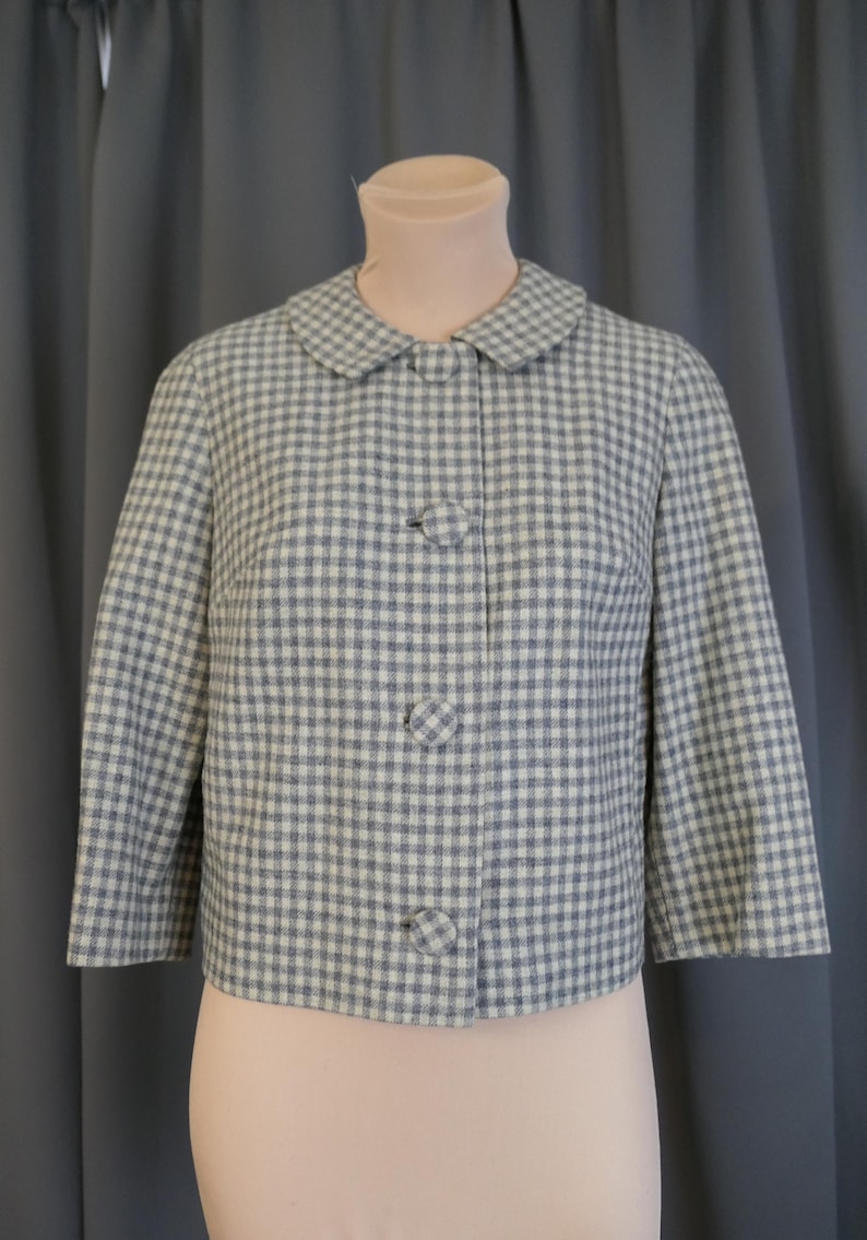 May include: A vintage-style jacket in a blue and white gingham pattern. The jacket features a rounded collar, three large matching buttons, and three-quarter sleeves. The jacket is displayed on a mannequin against a gray backdrop.