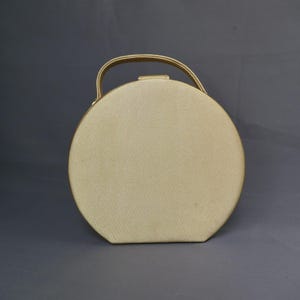 May include: A circular, cream-coloured handbag with a matching handle. The bag has a textured surface and a simple, elegant design. The handle is attached to the top of the bag.