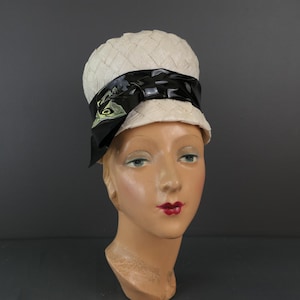 May include: A vintage cream-colored hat with a woven texture and a black satin ribbon. The ribbon is adorned with a green and black design. The hat is displayed on a mannequin head.