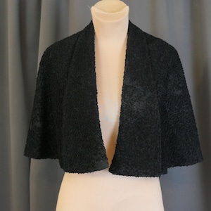 May include: A black, textured cape-style jacket with a wide, open front. The garment is displayed on a mannequin, showcasing its shape and design. The fabric appears to have a subtle pattern, adding visual interest to the overall look. The background is a neutral gray.
