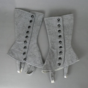 May include: A pair of grey spats with black buttons and a flared design. The spats have a strap that goes under the foot. The spats are made of a felt-like material.