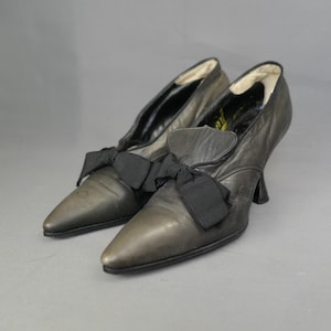 May include: A pair of vintage black leather pumps with pointed toes and small heels. Each shoe features a black bow detail on the vamp. The shoes are displayed on a gray background.