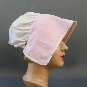 Può includere: Un cappello in stile vintage con un cappuccio bianco arricciato e una tesa rosa. Il bordo è rifinito con pizzo bianco. Il cappello è esposto su una testa di manichino su sfondo grigio.