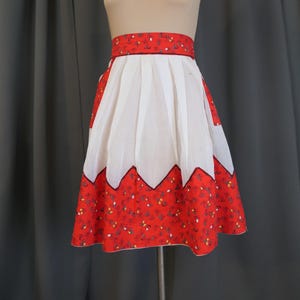 May include: A white apron with a red and white floral print border. The apron has a scalloped edge and a gathered waist.