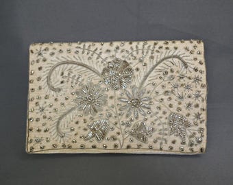 May include: Rectangular off-white clutch with intricate silver beaded floral embroidery. The design features three large flowers, leaves, and smaller star-shaped accents, all meticulously crafted with silver beads. The edges are also adorned with silver beads.