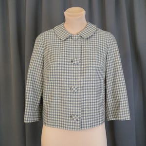May include: A vintage, short-sleeved jacket in a light blue and white gingham pattern. The jacket features a rounded collar and three matching buttons down the front. The garment is displayed on a mannequin.