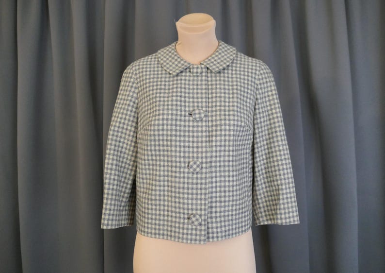 May include: A vintage, short-sleeved jacket in a light blue and white gingham pattern. The jacket features a rounded collar and three matching buttons down the front. The garment is displayed on a mannequin.