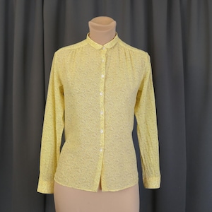 May include: A yellow long-sleeved blouse with a small floral pattern. The blouse has a button-down front and a rounded collar. The garment is displayed on a mannequin against a gray curtain backdrop.