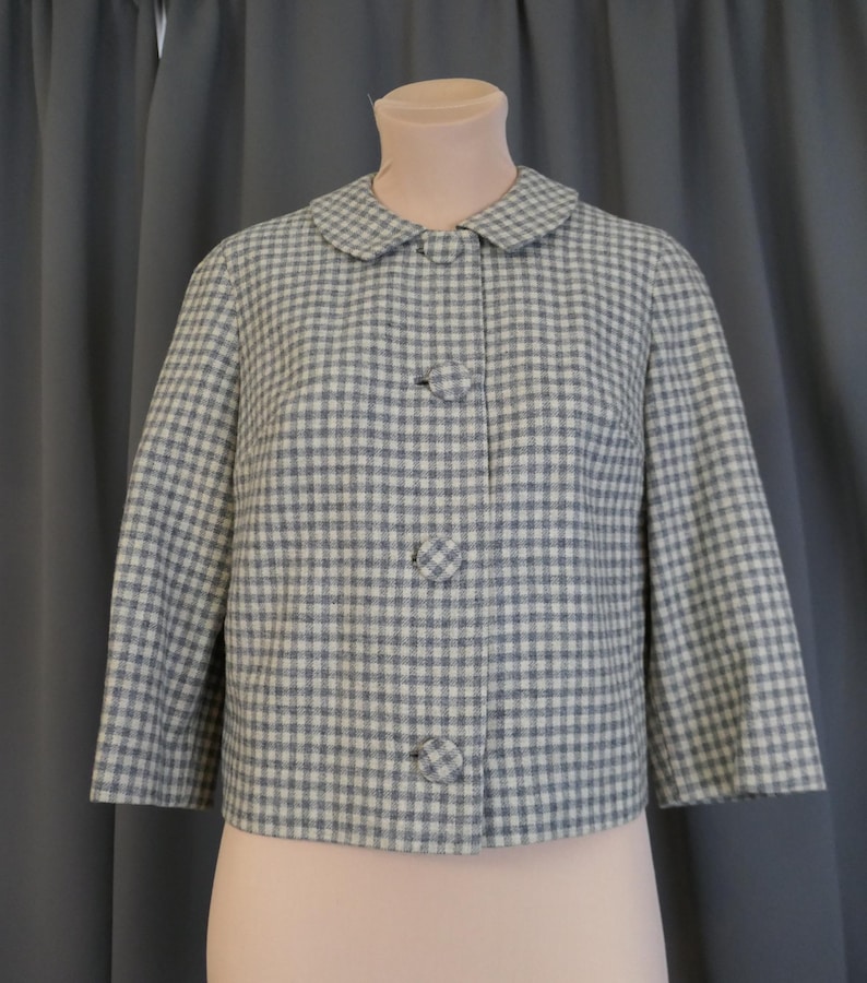 May include: A vintage-style jacket in a blue and white checkered pattern. The jacket has a rounded collar, three large matching buttons, and three-quarter sleeves. The fabric appears to be a woven material.