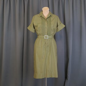 May include: Olive green dress with short sleeves, a collared neckline, and a button-down front. The dress features a matching belt with a silver-colored buckle. The dress is displayed on a mannequin against a gray curtain backdrop.