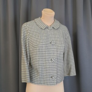 May include: A vintage cropped jacket in a blue and white gingham pattern. The jacket features a rounded collar, three decorative buttons, and three-quarter sleeves. The garment is displayed on a mannequin against a gray backdrop.