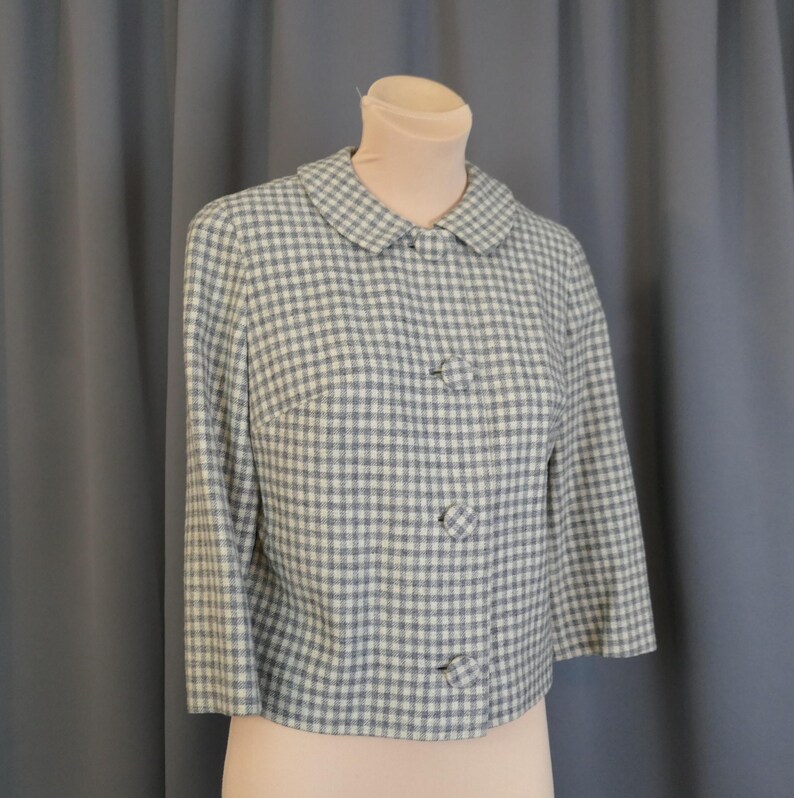 May include: A vintage cropped jacket in a blue and white gingham pattern. The jacket features a rounded collar, three decorative buttons, and three-quarter sleeves. The garment is displayed on a mannequin against a gray backdrop.