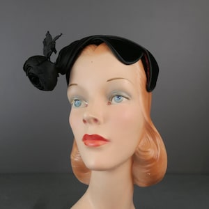 Puede incluir: Un sombrero negro con una copa lisa y curvada, adornado con una rosa de tela en el lateral. El sombrero está confeccionado con un material oscuro y sólido con un brillo sutil, diseñado para colocarse sobre la cabeza con una silueta redondeada y estructurada.