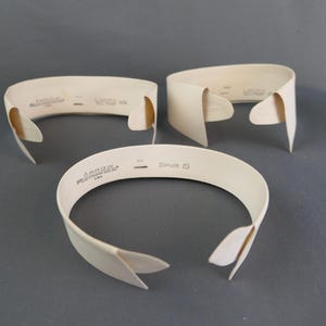 May include: Three off-white detachable shirt collars with pointed ends. Each collar features the printed text Arrow, Spur 15, and U.S.A. on the inner band. These stiff, curved garment accessories are arranged in a triangular formation on a flat surface.