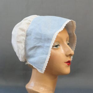 Könnte beinhalten: Eine Haube im Vintage-Stil, mit einer weißen Stoffbasis und einer hellblauen Krempe. Der Rand ist mit zarter weißer Spitze verziert. Die Haube wird auf einem Schaufensterkopf mit rotem Lippenstift präsentiert.