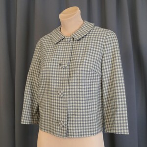 May include: A vintage, light blue and white checkered jacket with a round collar and three buttons. The jacket has three-quarter sleeves and is displayed on a mannequin. The fabric has a woven texture.