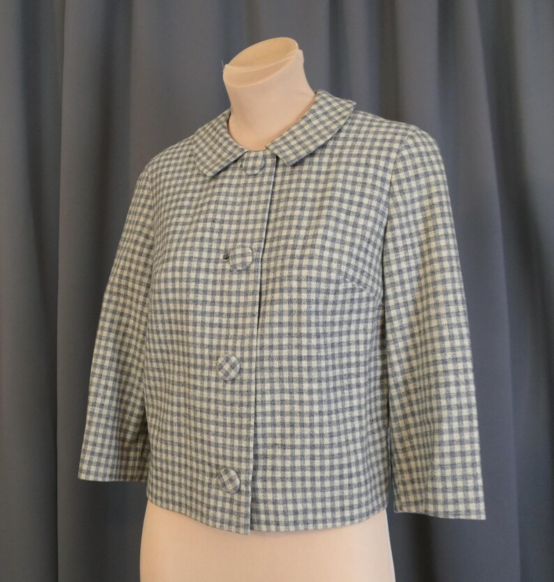 May include: A vintage, light blue and white checkered jacket with a round collar and three buttons. The jacket has three-quarter sleeves and is displayed on a mannequin. The fabric has a woven texture.