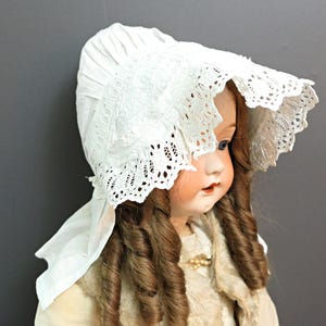 May include: A white lace bonnet with a ruffled edge. The bonnet is worn by a doll with brown curly hair.
