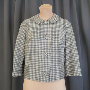 May include: A vintage, light blue and white checkered jacket with a rounded collar and three matching buttons. The jacket has three-quarter sleeves and is displayed on a mannequin. The fabric has a subtle texture.