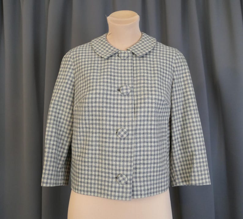 May include: A vintage, light blue and white checkered jacket with a rounded collar and three matching buttons. The jacket has three-quarter sleeves and is displayed on a mannequin. The fabric has a subtle texture.