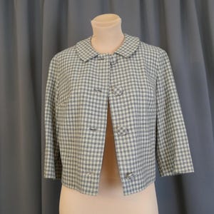 May include: A vintage cropped jacket in a blue and white gingham pattern. The jacket features a rounded collar, three-quarter sleeves, and three button closures. The garment is displayed on a mannequin against a neutral background.