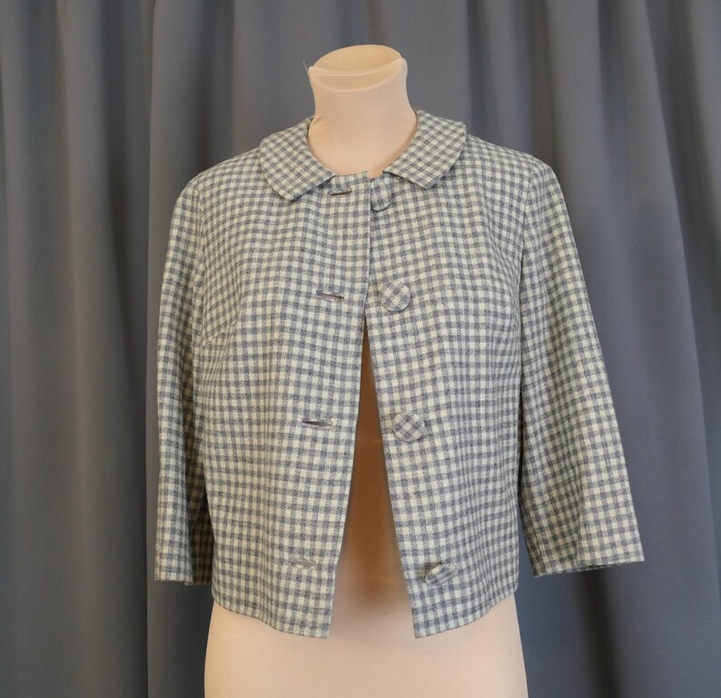 May include: A vintage cropped jacket in a blue and white gingham pattern. The jacket features a rounded collar, three-quarter sleeves, and three button closures. The garment is displayed on a mannequin against a neutral background.