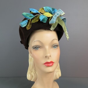 May include: A brown felt beret adorned with fabric leaves in shades of blue, green, and yellow. A light blue ribbon bow adds a decorative touch. The hat is displayed on a mannequin head with blonde hair and red lipstick.
