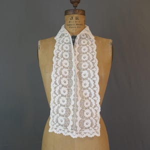 May include: A white lace collar featuring a repeating floral pattern with scalloped edges. The collar is displayed on a tan dress form. The lace has a delicate, openwork design with circular flower motifs arranged in vertical columns along the front.
