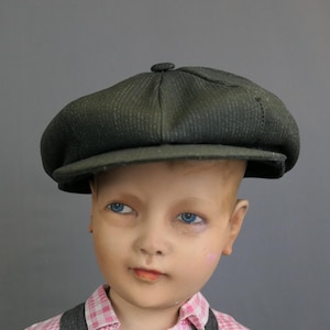 May include: A dark olive green newsboy cap with a subtle pinstripe pattern, featuring a rounded crown, a small top button, and a stiffened brim, worn on a mannequin head with blue eyes and a pink and white checkered collared shirt.