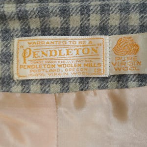 May include: Close-up of a Pendleton label, featuring text that reads "Warranted to be a Pendleton" and "100% Virgin Wool." The label is on a gray and beige plaid fabric, with a peach-colored lining visible.