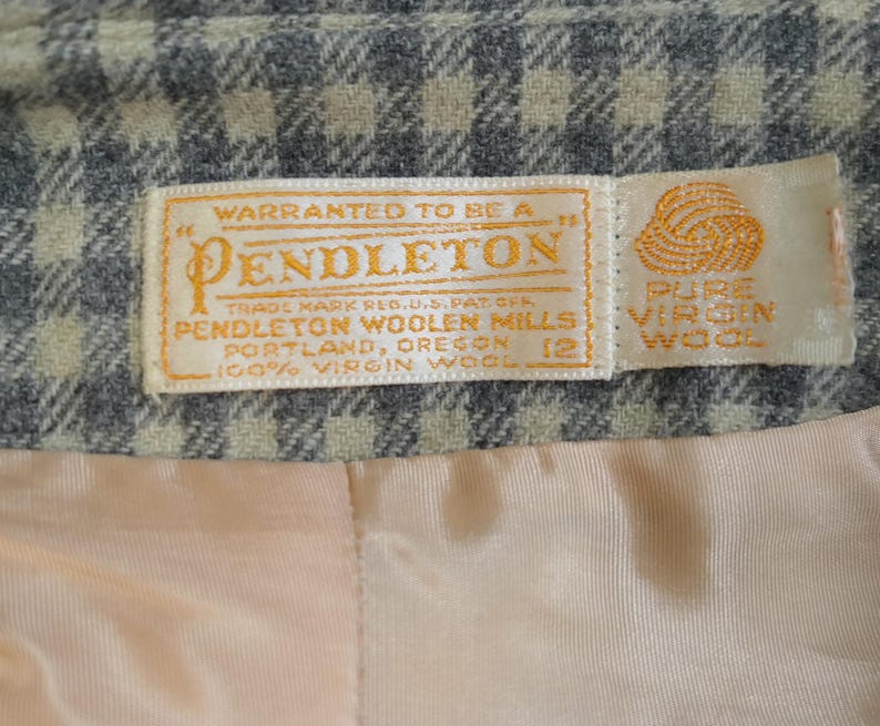May include: Close-up of a Pendleton label, featuring text that reads "Warranted to be a Pendleton" and "100% Virgin Wool." The label is on a gray and beige plaid fabric, with a peach-colored lining visible.