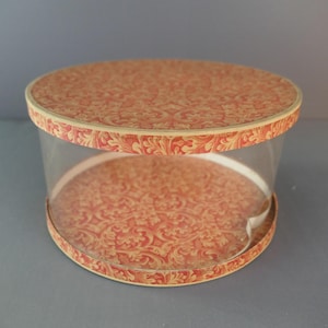 May include: A clear, round box with a lid and base featuring a red and beige floral pattern. The sides of the box are transparent, allowing a view of the contents. The box is designed for storage or display.