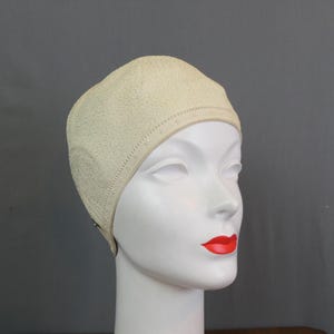 May include: A cream-colored swim cap with a textured, pebbled surface and a decorative band featuring small, repeating geometric cutouts along the lower edge. The cap is fitted snugly over the head of a white mannequin with red painted lips.