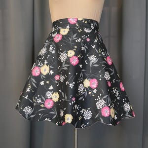 May include: A black flared skirt with a floral pattern. The skirt features pink, yellow, and white flowers with green leaves. The skirt is displayed on a mannequin and has a scalloped hemline.