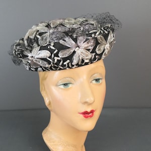 May include: A vintage black hat with white and silver floral embroidery and a black mesh veil. The hat is displayed on a mannequin head with red lipstick and blue eye shadow. The hat is a stylish accessory.