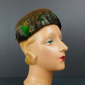 Puede incluir: Un sombrero de fieltro marrón con una banda de plumas verdes y marrones.