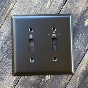 May include: A black metal light switch plate with two toggle switches. The top switch has a light bulb icon and the bottom switch has a ceiling fan icon.