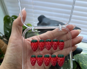 May include: A clear plastic box with a lid containing 10 red and green strawberry-shaped nail tips.